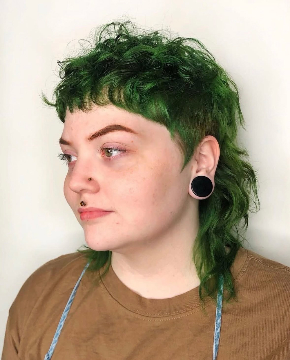 Side profile of a person with a short, textured green mohawk, pink ear plugs, nose piercings, wearing a brown top, looking to the left.