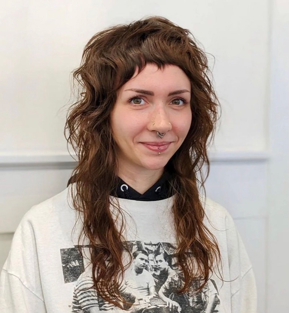 Portrait of a young woman with wavy brown hair and facial piercings, wearing a white graphic hoodie and smiling at the camera.