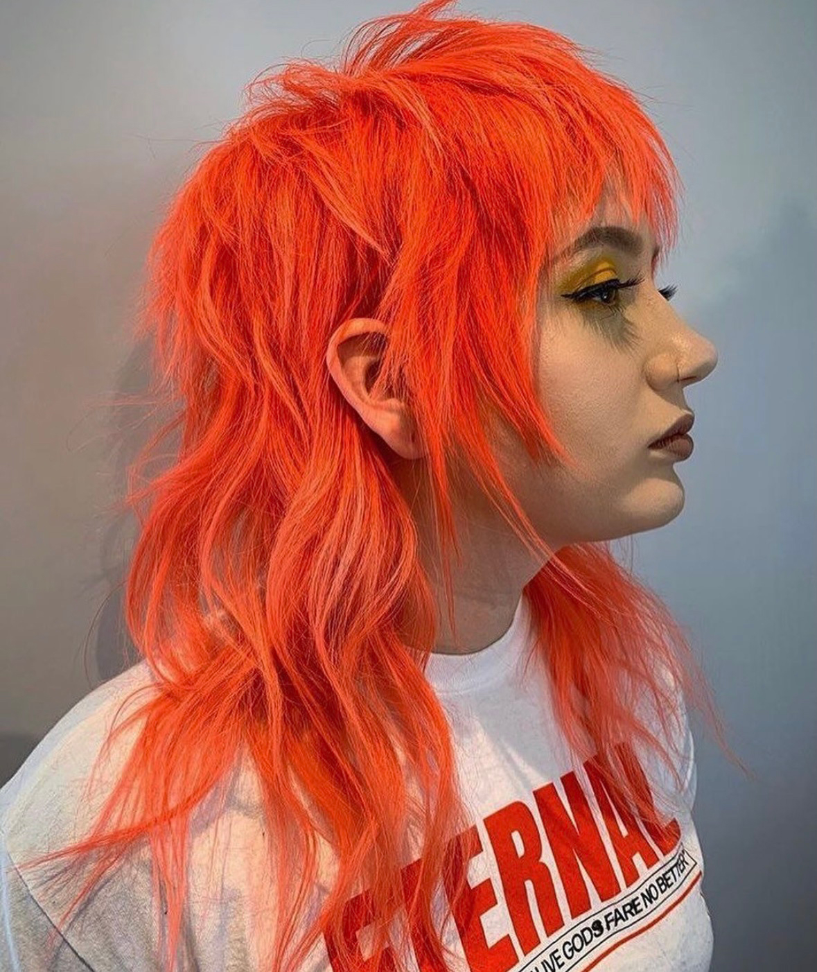 Side profile of a person with neon orange, spiky mullet hair and bold winged eyeliner, wearing a white shirt with red text.