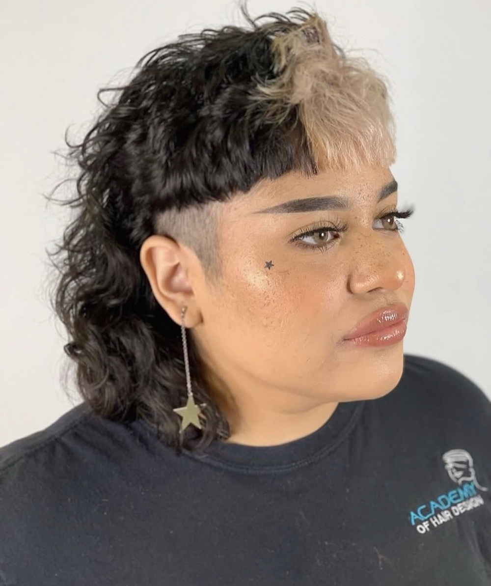 Portrait of a person with a black-and-blonde mullet, side shave, star cheek tattoo, and dangling star earring wearing a black shirt with a logo on the chest area.
