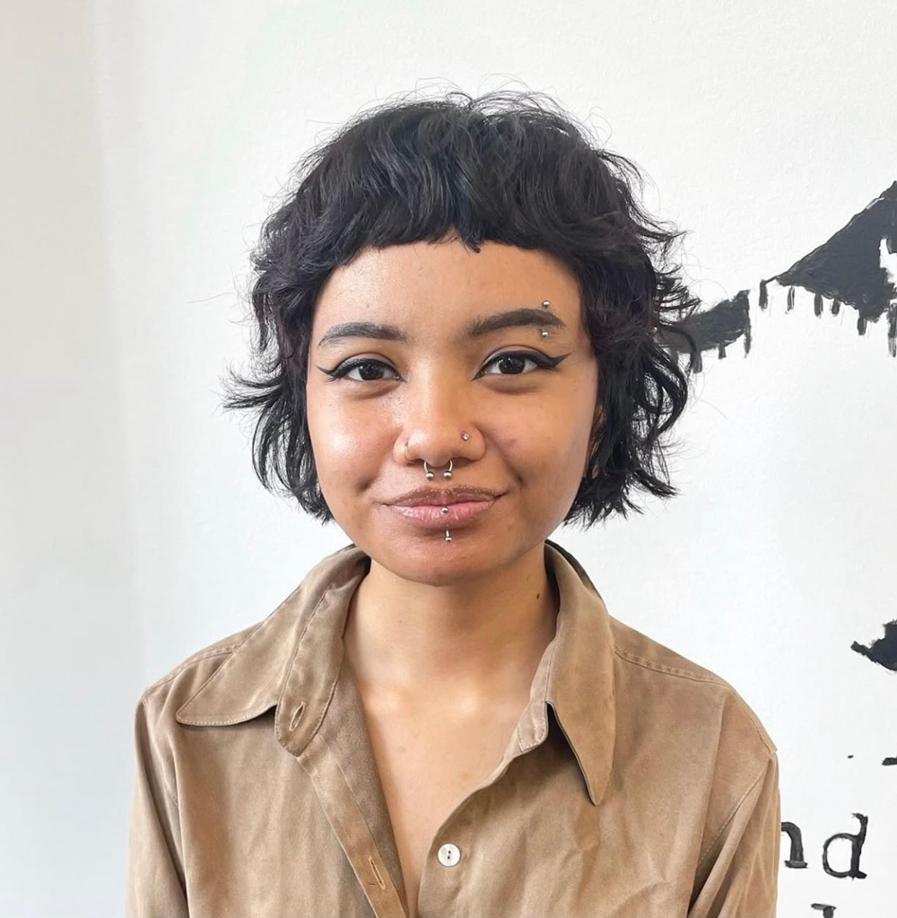 Portrait of a person with short black wavy hair, winged eyeliner, and multiple facial piercings, wearing a brown button-down shirt.