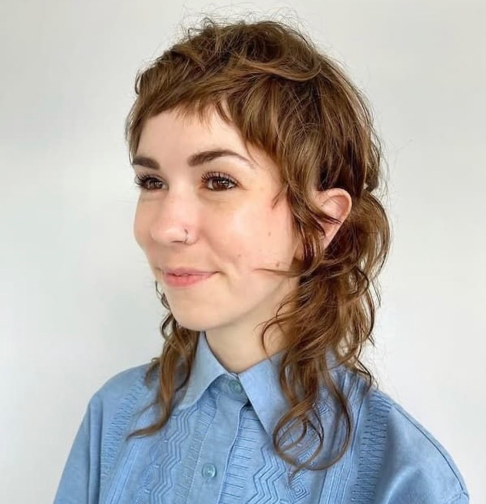 Portrait of a woman with short, wavy brown hair and a nose ring, wearing a light blue button-down shirt, looking to the left.