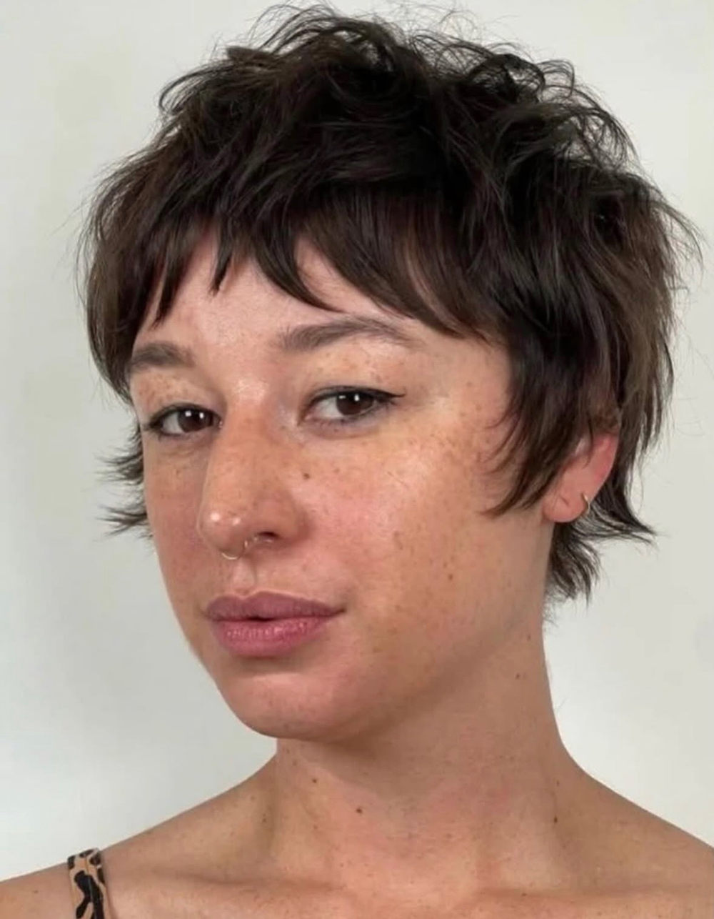 Close-up portrait of a person with short messy brown hair, freckles, and a small nose hoop, looking slightly to the side.