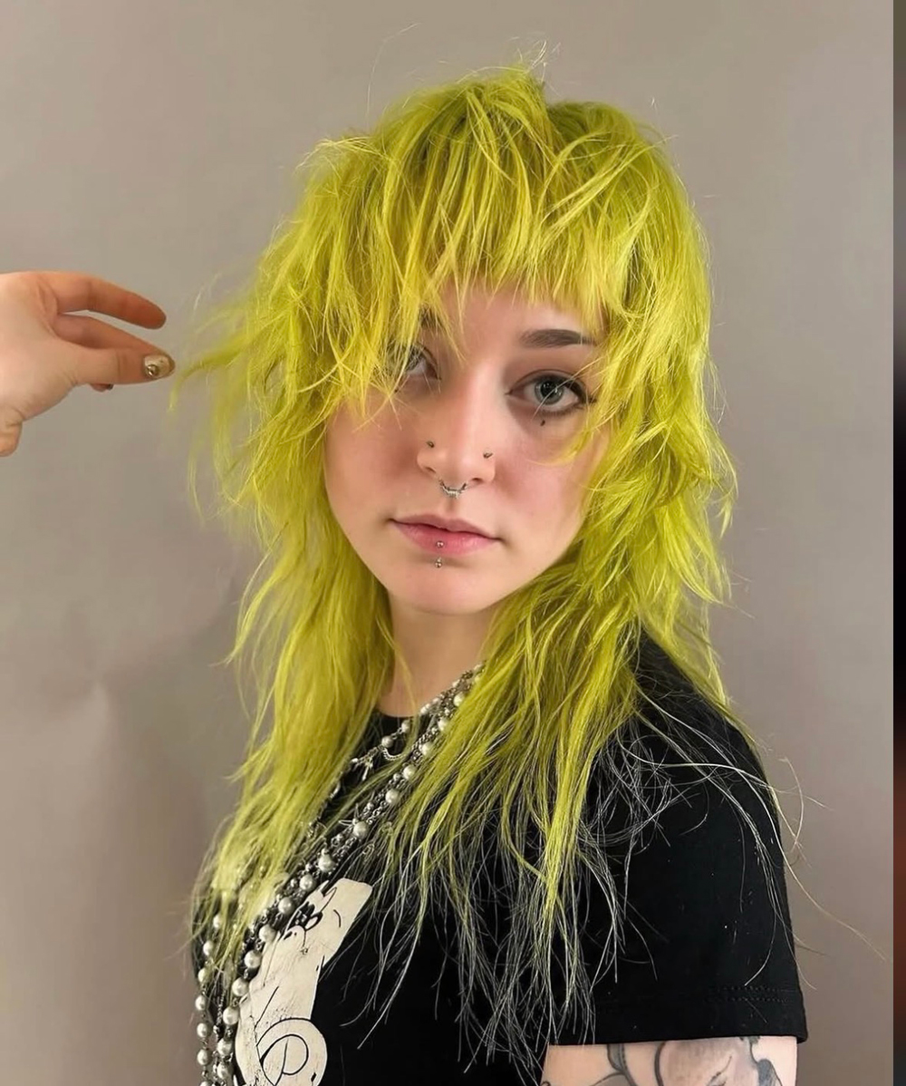 Person with neon yellow, shaggy hairstyle and facial piercings (septum and lip) wearing a black graphic tee and layered necklaces, posing to the side.